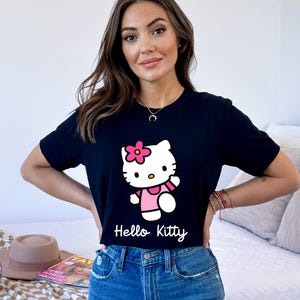 May include: Black t-shirt featuring a Hello Kitty graphic. The graphic shows Hello Kitty in a pink dress with a pink flower on her head. The words "Hello Kitty" are written in white script below the graphic.