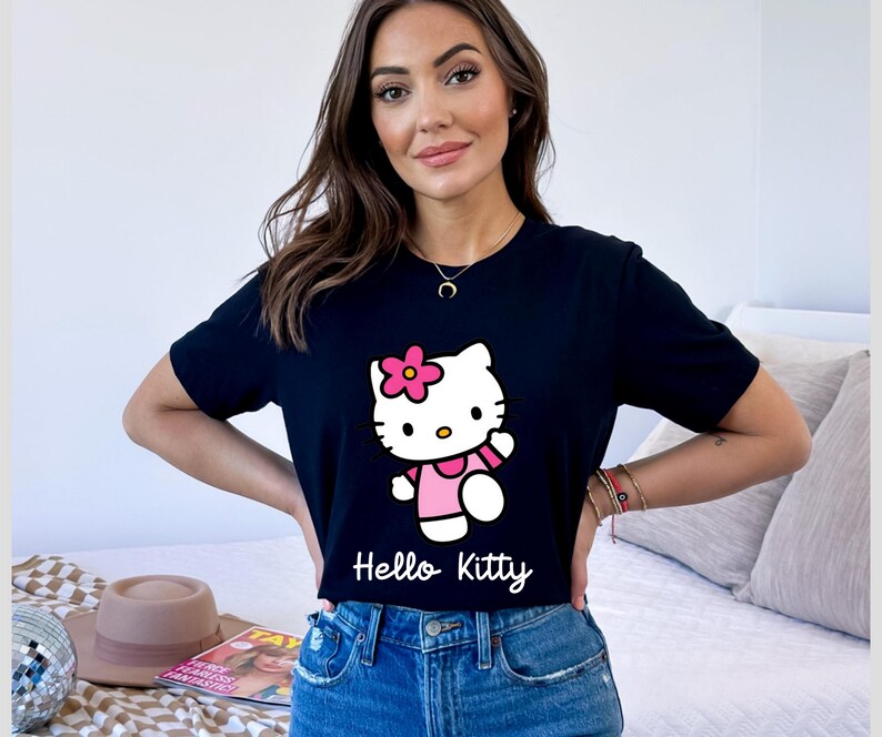 May include: Black t-shirt featuring a Hello Kitty graphic. The graphic shows Hello Kitty in a pink dress with a pink flower on her head. The words "Hello Kitty" are written in white script below the graphic.