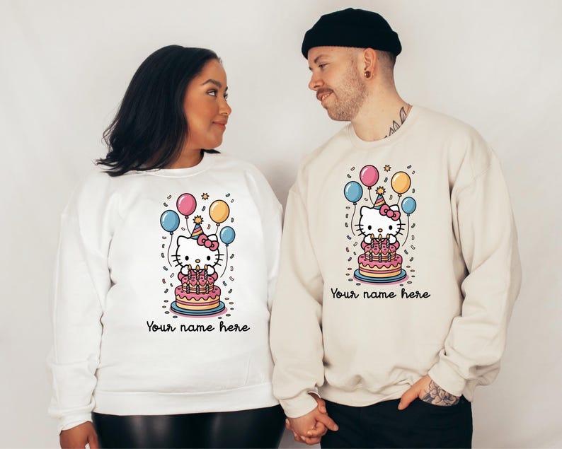May include: White and beige sweatshirts featuring a Hello Kitty birthday design. The design includes Hello Kitty on a cake with balloons and the text "Your name here". The sweatshirts are a light color.
