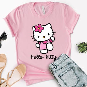 May include: A pink t-shirt featuring a Hello Kitty graphic. The graphic shows Hello Kitty wearing a pink dress and a flower on her head. The words "Hello Kitty" are printed below the graphic. The shirt is paired with blue denim shorts, sunglasses, and sandals.