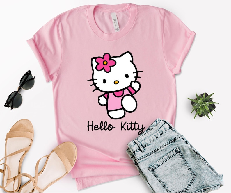 May include: A pink t-shirt featuring a Hello Kitty graphic. The graphic shows Hello Kitty wearing a pink dress and a flower on her head. The words "Hello Kitty" are printed below the graphic. The shirt is paired with blue denim shorts, sunglasses, and sandals.