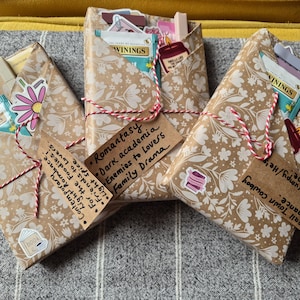 Könnte beinhalten: Drei Geschenkpakete in floral gemustertem Papier, gebunden mit rot-weißem Garn. Jedes hat ein Etikett mit handschriftlichem Text. Aufkleber und Teebeutel sind sichtbar. Die Etiketten erwähnen Themen wie "Romantasy" und "Small Town Cowboy".