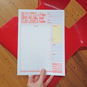 May include: A white paper habit tracker sheet titled Silly Little Star Charts 75-Day Habit Tracker in red text. It features a grid of seventy-five numbered stars, sections for daily habits, a why statement, rewards, and a report card at the bottom.