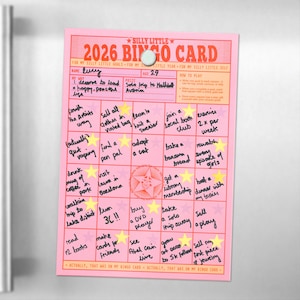 Könnte beinhalten: Eine rosa 2026 Bingo-Karte mit handgeschriebenen Zielen und Sternaufklebern, die mit einem Magneten an einem Kühlschrank befestigt ist. Die Karte enthält Ziele wie 'einem lokalen Buchclub beitreten' und '2x pro Woche trainieren'.