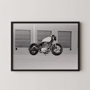 Könnte beinhalten: Schwarz-Weiß-Druck in einem Rahmen eines Custom-Motorrads. Das Motorrad hat einen weißen Kraftstofftank, schwarze Reifen und ein minimalistisches Design. Der Hintergrund zeigt ein Gebäude mit drei Garagentoren.
