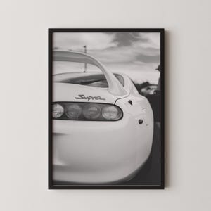 Könnte beinhalten: Schwarz-Weiß-Druck eines weißen Supra-Sportwagens in einem Rahmen. Das Heck des Autos ist zu sehen, mit Rückleuchten, Spoiler und dem "Supra"-Logo. Der Hintergrund zeigt einen bewölkten Himmel.