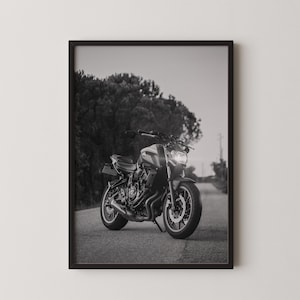 May include: Black and white framed photograph of a motorcycle parked on a paved road. The motorcycle is facing the viewer, with its headlights illuminated. The background features a blurred view of trees and a road.