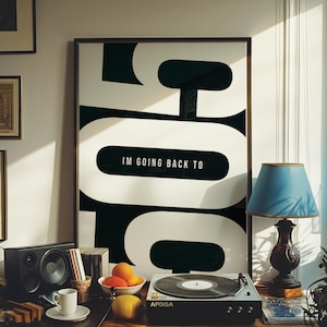 May include: A framed black and white poster with the text "IM GOING BACK TO" is displayed above a record player, speaker, and fruit. A blue lampshade sits on a dark brown lamp. A white coffee cup and saucer are on the table.