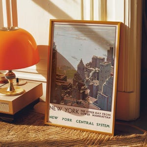 May include: A framed vintage travel poster of New York City, featuring a cityscape view from the Upper Bay. The poster has a gold frame and includes the text "New York Central System". An orange lamp sits to the left.