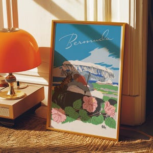 May include: A framed travel poster with the word "Bermuda" in script. The poster features a beach scene with a couple under a white umbrella, pink hibiscus flowers, and a blue sky. The frame is gold.