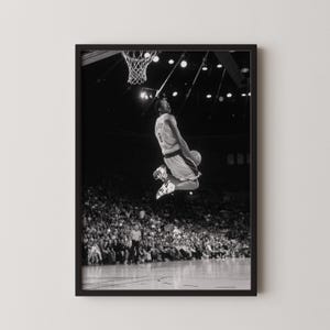 May include: Monochrome framed print showing a basketball player in mid-air, poised to dunk. The player wears a jersey with the number 8. The background reveals a packed stadium and the basketball hoop.
