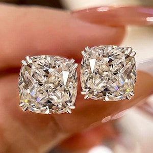 May include: A pair of square-cut diamond stud earrings. The diamonds are clear and sparkling, set in a silver or white gold setting. The earrings are shown close up, highlighting their brilliance and design.