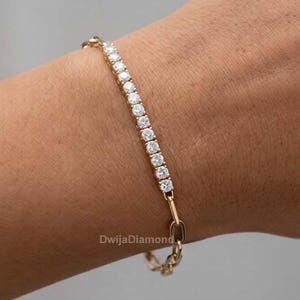 May include: A gold bracelet featuring a line of clear, round gemstones. The bracelet is worn on a wrist, with a small chain link clasp. This delicate piece of jewellery is a stylish accessory.