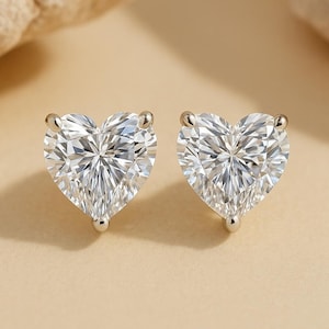 May include: A pair of heart-shaped stud earrings. Each earring features a clear, faceted gemstone set in a silver-colored metal. The gemstones are cut to maximize brilliance and sparkle. The earrings are displayed on a neutral, light-colored background.