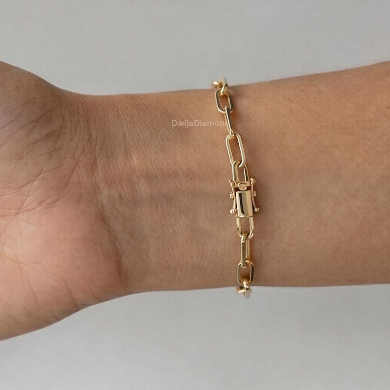 May include: A gold-coloured link bracelet with a rectangular clasp is shown on a wrist. The bracelet is made of elongated, rectangular links. The clasp is centred and has a rectangular shape. The bracelet is on a light skin tone.