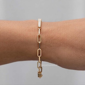 May include: A gold-coloured link bracelet with a row of small, clear stones at the top. The bracelet is on a person's wrist. The bracelet has a rectangular clasp at the bottom. The background is a neutral colour.