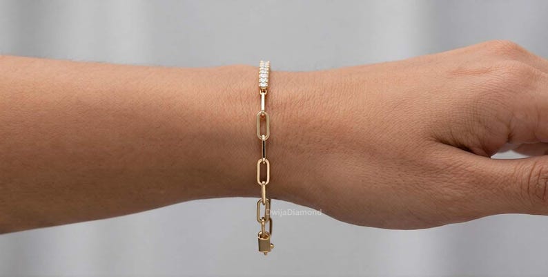 May include: A gold-coloured link bracelet with a row of small, clear stones at the top. The bracelet is on a person's wrist. The bracelet has a rectangular clasp at the bottom. The background is a neutral colour.