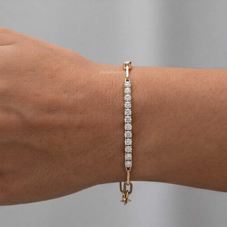 May include: A gold bracelet featuring a line of small, clear gemstones. The bracelet is fastened with a clasp. The bracelet is on a person's arm.