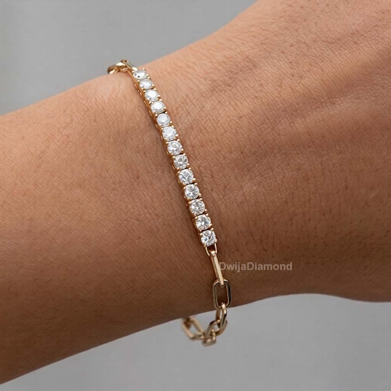 May include: A gold bracelet with a line of clear, round gemstones. The bracelet has a delicate design with a chain-link clasp. The gemstones are set in a gold band, creating a sparkling and elegant accessory.