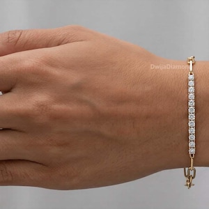 May include: A gold bracelet featuring a line of small, clear gemstones. The bracelet is fastened with a clasp at the bottom and a small decorative element at the top. The bracelet is displayed on an arm.