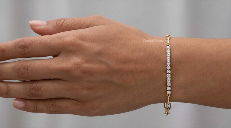 May include: A gold bracelet featuring a line of small, clear gemstones. The bracelet is fastened with a clasp at the bottom and a small decorative element at the top. The bracelet is displayed on an arm.