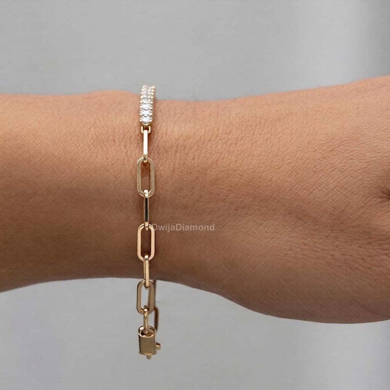 May include: A gold-coloured link bracelet featuring a row of small, clear stones at the top. The bracelet is made up of elongated oval links and is fastened with a clasp. The bracelet is shown on a person's wrist.