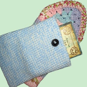 May include: A light blue and white crocheted case with a black button closure. The case holds a book with the text "The New York Times Bestselling Series Michael Scott". A colorful crocheted coaster is visible behind the case.