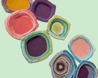 May include: A collection of handmade crochet baskets in various colors, including yellow, pink, purple, and blue. The baskets have a square or hexagonal shape with a textured stitch pattern. The image is taken from a slightly elevated angle.