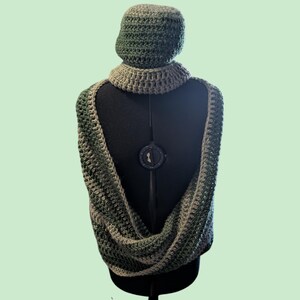 Op de afbeelding: Een handgemaakte haakset in groen- en grijstinten, bestaande uit een muts, col en infinity sjaal. De set wordt getoond op een zwarte mannequin, waarbij de textuur en het ontwerp van het garen worden benadrukt. Ideaal voor de winter.