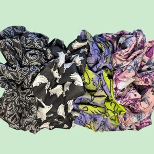 Puede incluir: Cuatro scrunchies de tela con diferentes patrones. Uno es negro con siluetas de pájaros blancos, otro es verde con flores moradas, uno es morado con flores verdes y otro es rosa con flores moradas.