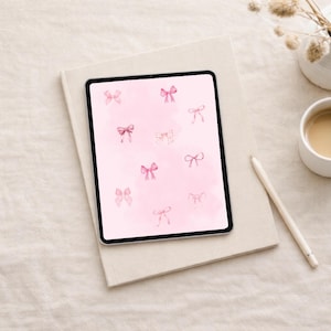 Pode incluir: Um tablet digital exibe um fundo rosa em aquarela com laços rosa espalhados. O tablet está sobre um livro de cor clara, ao lado de uma xícara de café, um lápis e um pequeno vaso com flores secas. A cena é montada em uma superfície de tons neutros.