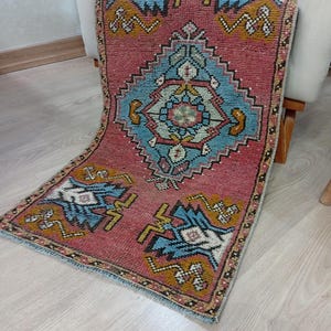May include: A small, rectangular rug with a faded red background and intricate geometric patterns in shades of blue, yellow, and white. The rug has a central diamond-shaped design and borders with repeating motifs. The rug is placed on a light-colored wooden floor.