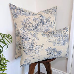 Peut inclure: Deux coussins décoratifs avec un motif toile de Jouy bleu et crème. Le tissu présente des scènes pastorales détaillées avec des personnages, des arbres et du feuillage. Un coussin est carré et l'autre rectangulaire, avec le même imprimé.