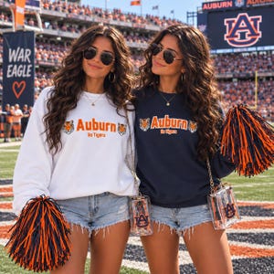 May include: Two long-sleeve crewneck sweatshirts, one white and one navy blue, both featuring the text Auburn Go Tigers and tiger head graphics. Each sweatshirt is paired with light blue denim shorts, sunglasses, and a clear crossbody bag.