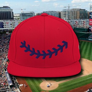 May include: A red baseball cap with a navy blue stitched baseball seam design on the front. The cap is in front of a baseball stadium.