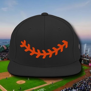May include: A black baseball cap with an orange stitched baseball seam design on the front. The cap is sitting on a baseball field.