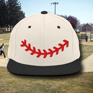 May include: A white baseball cap with a black brim. The front of the cap has a red baseball stitch design that forms a smiley face.