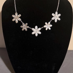 Może przedstawiać: Srebrny zestaw biżuterii w kwiaty, w tym naszyjnik, bransoletka i kolczyki, prezentowany na czarnym manekinie. Naszyjnik ma pięć kwiat&oacute;w przypominających stokrotki. Bransoletka znajduje się na g&oacute;rze manekina.