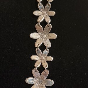 Może przedstawiać: Srebrna bransoletka z zawieszkami w kształcie stokrotek. Każda zawieszka ma szczeg&oacute;łowe wzory płatk&oacute;w i środka. Bransoletka ma przedłużenie łańcuszka i zawieszkę w kształcie serca na końcu.