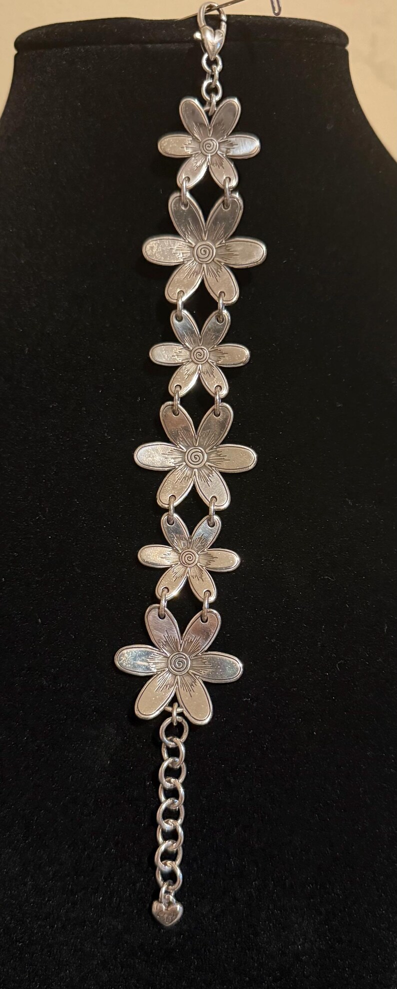 Może przedstawiać: Srebrna bransoletka z zawieszkami w kształcie stokrotek. Każda zawieszka ma szczeg&oacute;łowe wzory płatk&oacute;w i środka. Bransoletka ma przedłużenie łańcuszka i zawieszkę w kształcie serca na końcu.