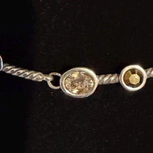 May include: Silver-toned necklace with three decorative elements. The necklace features a twisted rope design. The decorative elements include a round pearl, an oval-shaped light brown gemstone, and a round, gold-colored gemstone.