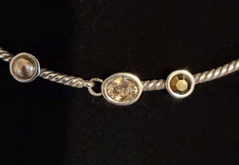 May include: Silver-toned necklace with three decorative elements. The necklace features a twisted rope design. The decorative elements include a round pearl, an oval-shaped light brown gemstone, and a round, gold-colored gemstone.
