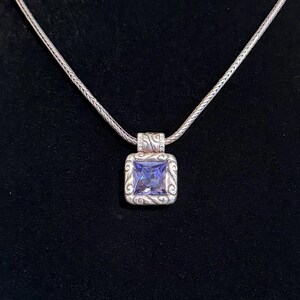 Puede incluir: Un collar de plata con un colgante cuadrado con una gran piedra preciosa de color p&uacute;rpura claro. El colgante est&aacute; enmarcado por un dise&ntilde;o ornamental y arremolinado. La cadena es de un estilo delicado y tejido, y el fondo es negro.