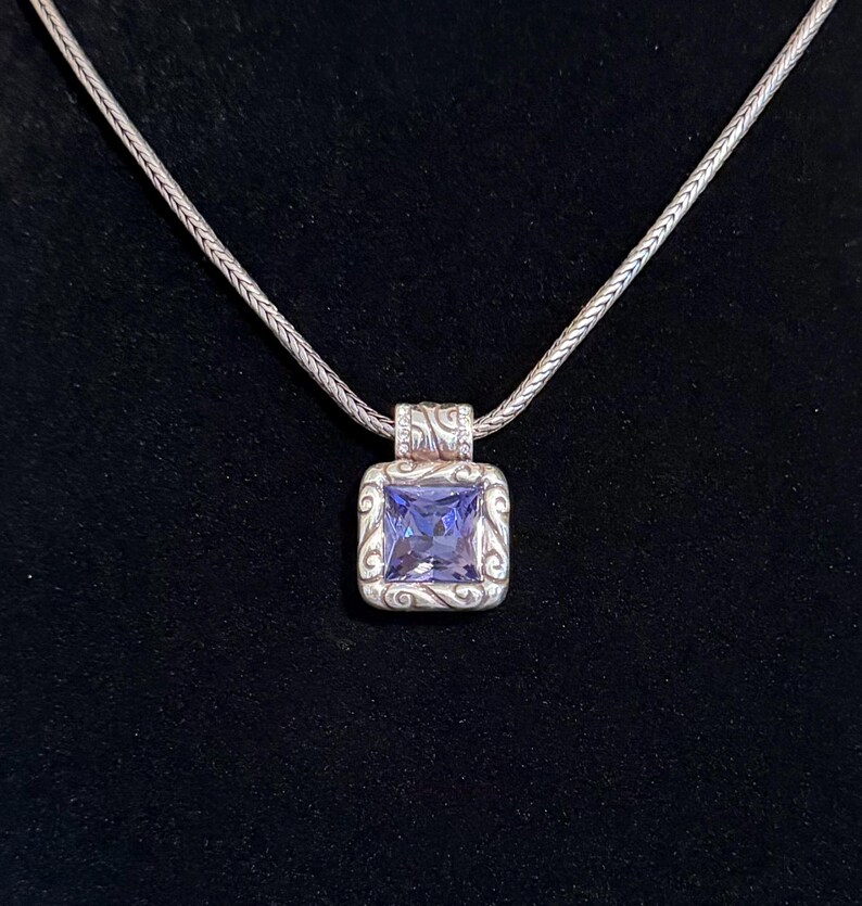Puede incluir: Un collar de plata con un colgante cuadrado con una gran piedra preciosa de color p&uacute;rpura claro. El colgante est&aacute; enmarcado por un dise&ntilde;o ornamental y arremolinado. La cadena es de un estilo delicado y tejido, y el fondo es negro.