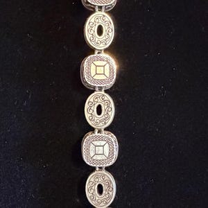 May include: Silver bracelet with alternating square and oval links. Each link has an ornate design with a central gold-coloured square or circle. The bracelet is shown against a dark background.