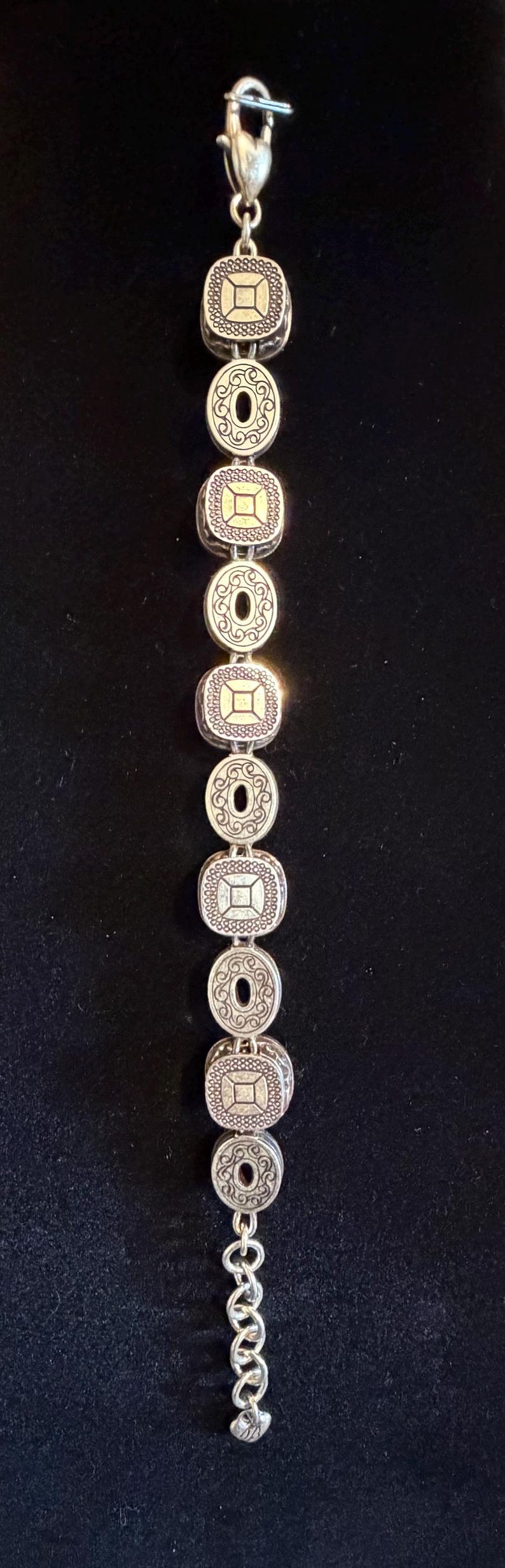 May include: Silver bracelet with alternating square and oval links. Each link has an ornate design with a central gold-coloured square or circle. The bracelet is shown against a dark background.
