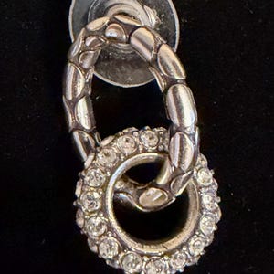 Puede incluir: Pendiente plateado con un dise&ntilde;o de doble bucle. El bucle superior tiene un patr&oacute;n texturizado y acanalado, mientras que el bucle inferior est&aacute; incrustado con peque&ntilde;os cristales brillantes. El poste del pendiente es un disco plateado.