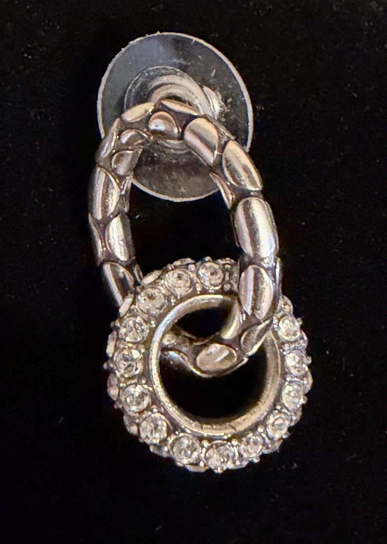 Puede incluir: Pendiente plateado con un dise&ntilde;o de doble bucle. El bucle superior tiene un patr&oacute;n texturizado y acanalado, mientras que el bucle inferior est&aacute; incrustado con peque&ntilde;os cristales brillantes. El poste del pendiente es un disco plateado.