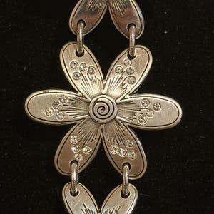 Może przedstawiać: Srebrna bransoletka z motywem kwiatowym z trzema wisiorkami w kształcie kwiat&oacute;w. Każdy kwiat ma spiralny środek i detale płatk&oacute;w. Płatki zdobią małe, przezroczyste kryształki. Bransoletka połączona jest małymi, zakrzywionymi ogniwami.