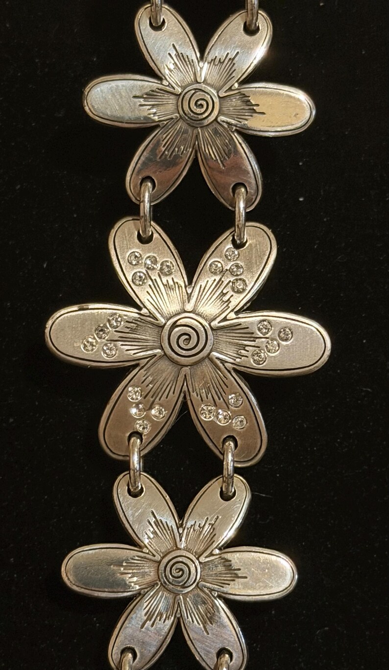 Może przedstawiać: Srebrna bransoletka z motywem kwiatowym z trzema wisiorkami w kształcie kwiat&oacute;w. Każdy kwiat ma spiralny środek i detale płatk&oacute;w. Płatki zdobią małe, przezroczyste kryształki. Bransoletka połączona jest małymi, zakrzywionymi ogniwami.